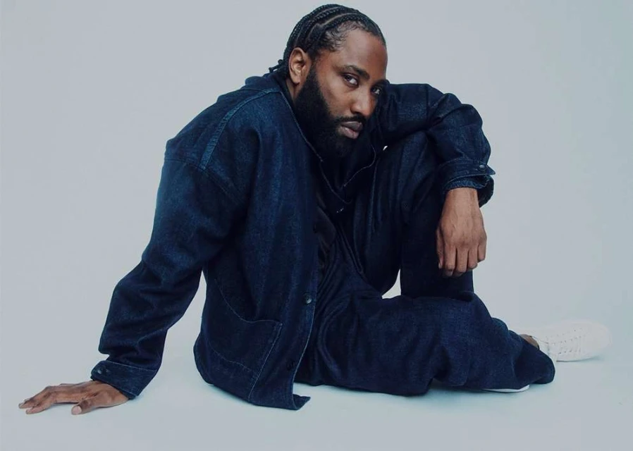 Portrait of a stylish Black male model with braided hair, posing in a denim jacket and pants on a light background in a studio setting.