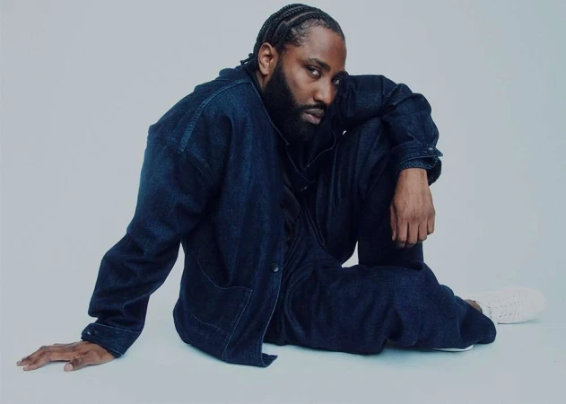 Portrait of a stylish Black male model with braided hair, posing in a denim jacket and pants on a light background in a studio setting.