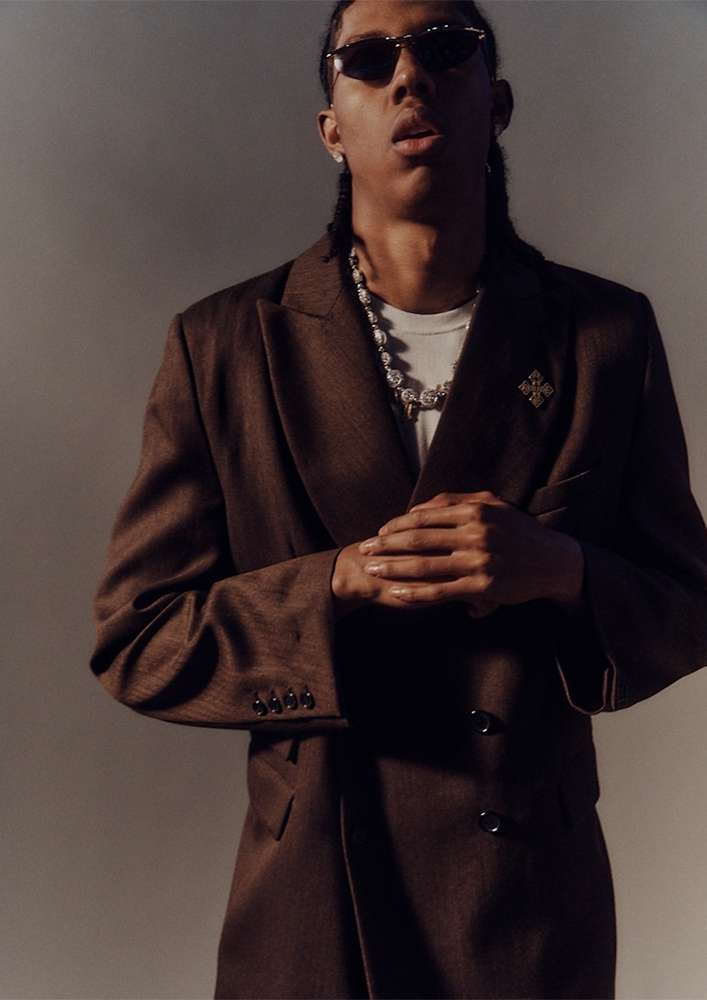Portrait of a man wearing sunglasses, a brown blazer, a white t-shirt, and a silver chain necklace against a grey background.