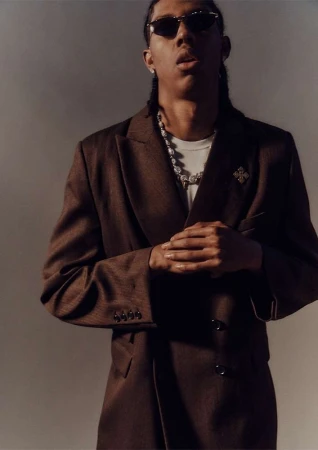 Portrait of a man wearing sunglasses, a brown blazer, a white t-shirt, and a silver chain necklace against a grey background.