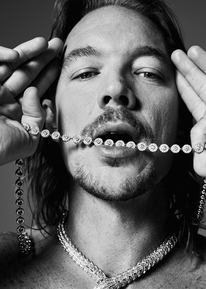 Monochrome portrait of a shirtless man with a goatee, holding a diamond necklace in his mouth and wearing a second necklace.