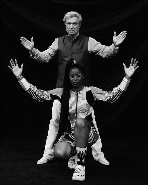 Two people in a studio pose with arms raised, showcasing fashion and style in a black and white photograph with high contrast.