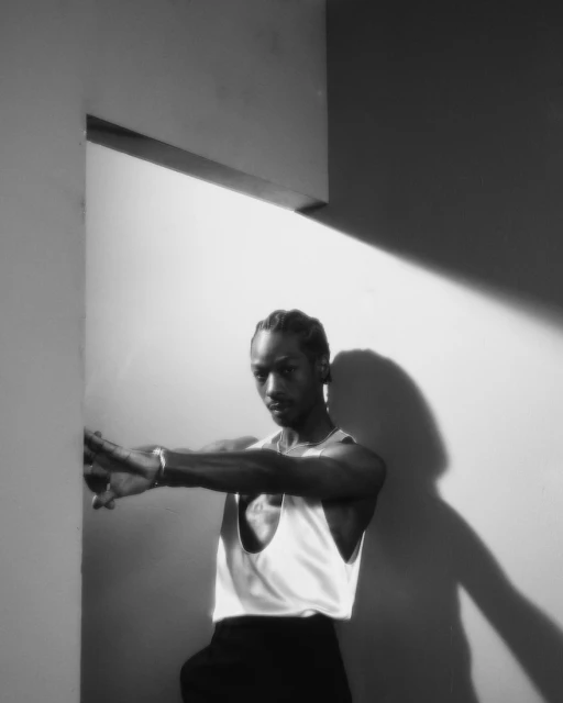 Black and white studio portrait of a Black adult male wearing a tank top, with a striking shadow cast on the wall behind him.