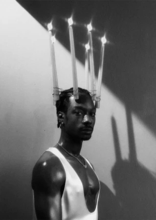 Black man wearing a crown of lit candles and a white tank top against a wall with harsh diagonal shadows.