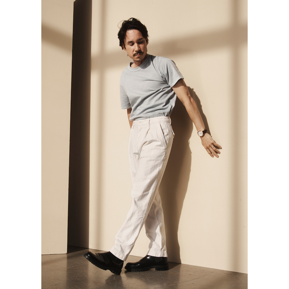 An adult male poses in a photo studio wearing a light blue t-shirt, white dress pants, and black dress shoes. Shadow patterns on the wall.