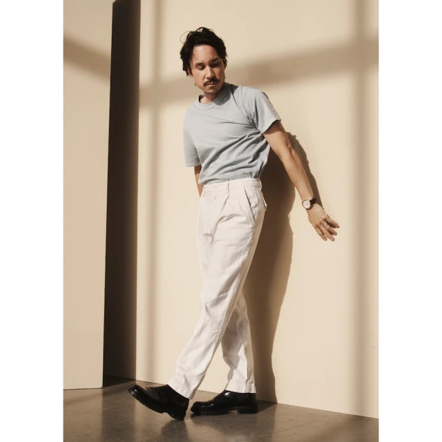 An adult male poses in a photo studio wearing a light blue t-shirt, white dress pants, and black dress shoes. Shadow patterns on the wall.