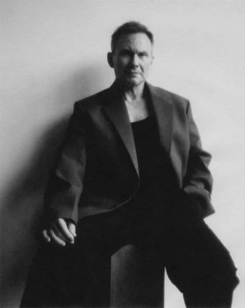 Monochrome portrait of an adult man in a blazer sitting against a plain white wall by Erik Tanner