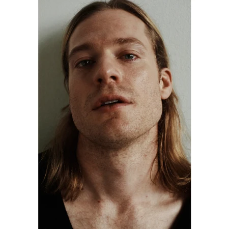 Close-up portrait of a man with long blond hair and stubble against a neutral wall by Erik Tanner