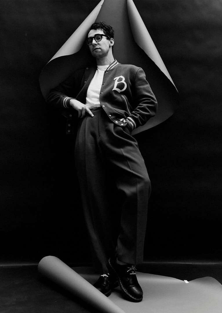Stylish man in letterman jacket poses against a black backdrop with rolled paper. He wears glasses, tailored pants, and dress shoes.