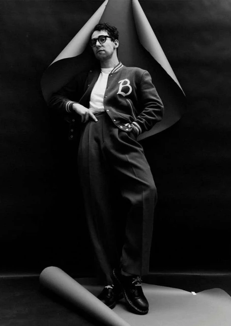 Stylish man in letterman jacket poses against a black backdrop with rolled paper. He wears glasses, tailored pants, and dress shoes.