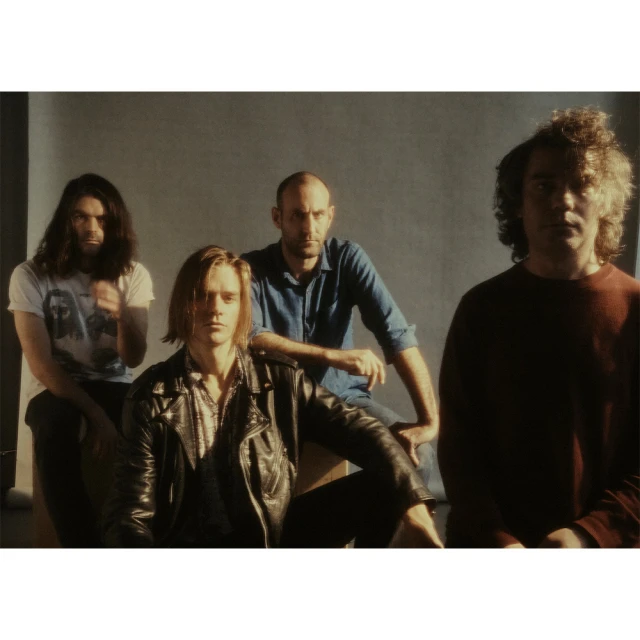 A group of four casually dressed adult men sit in a dimly lit indoor setting, posing for a band portrait with a serious expression.