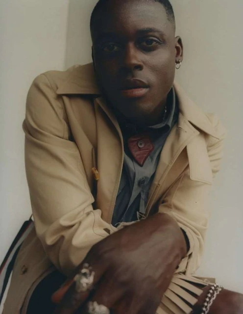 Portrait of a stylish young man with dark skin wearing a tan trench coat and jewelry indoors against a white background.