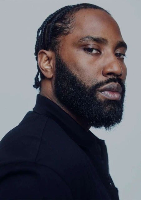Portrait of a black man with a beard and braided hair wearing a black shirt against a grey background, looking at the camera.