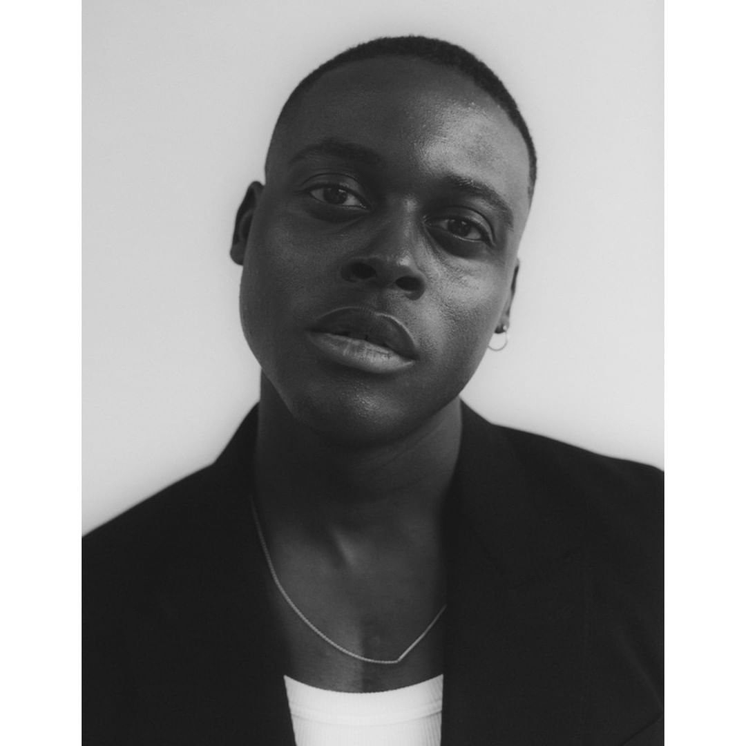 Monochrome portrait of a black man wearing a dark jacket and a necklace against a plain light background