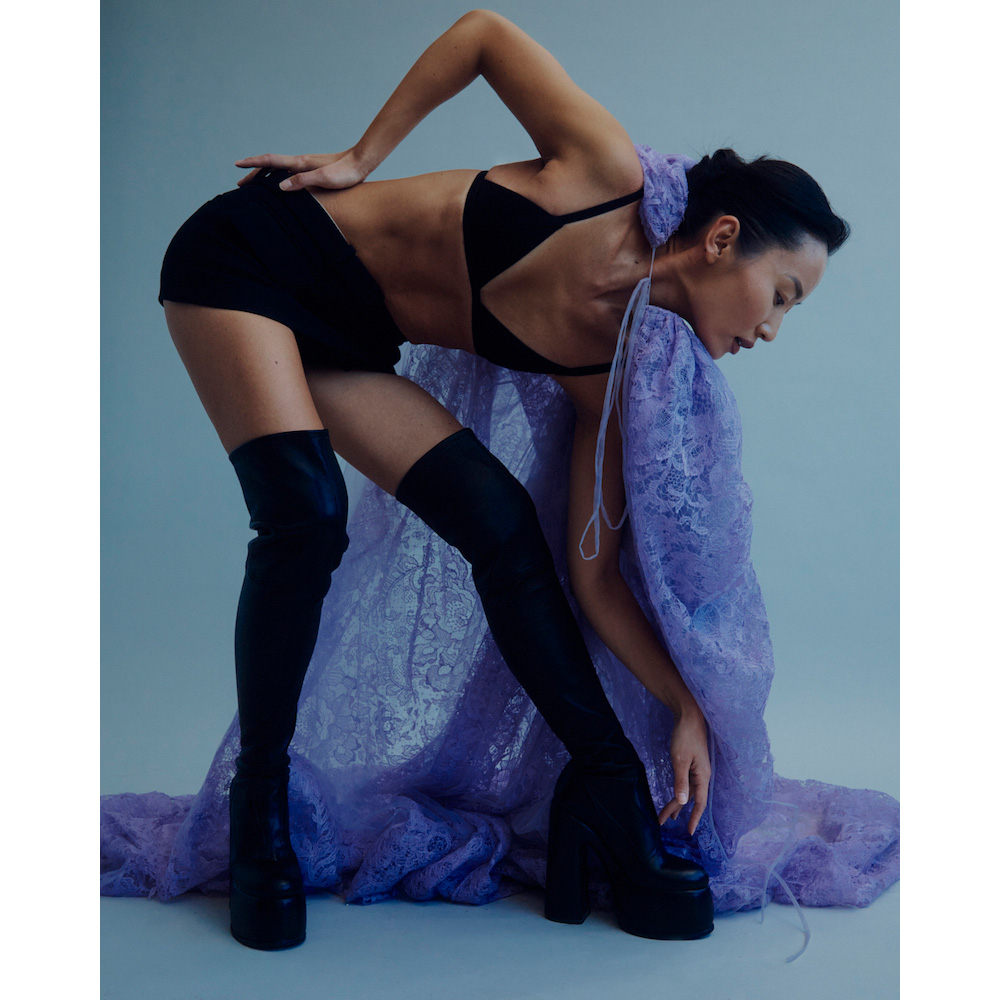 Woman poses in black bra, shorts, thigh-high boots, and a sheer lavender lace shawl in a studio setting with soft lighting.