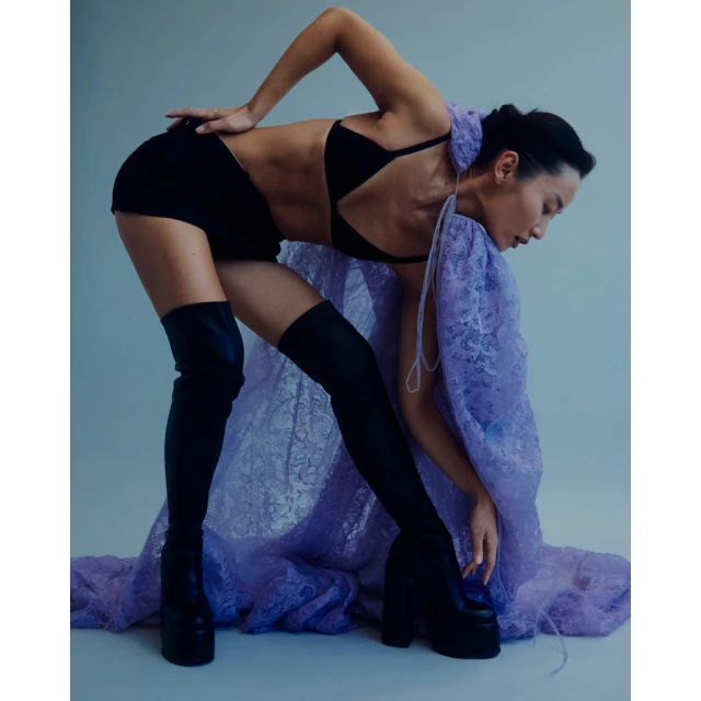 Woman poses in black bra, shorts, thigh-high boots, and a sheer lavender lace shawl in a studio setting with soft lighting.