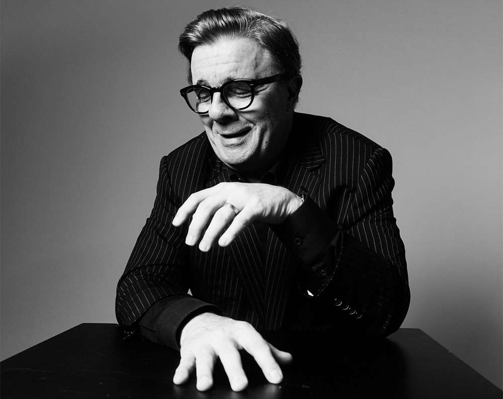 Monochrome portrait of an older man in a pinstripe suit and glasses gesturing with his hands over a dark table.