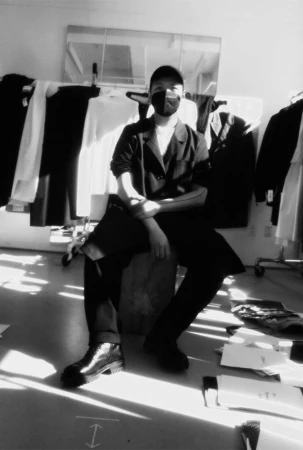 Monochrome portrait of a seated person wearing a black mask, cap, and dark jacket in a studio with clothing racks.