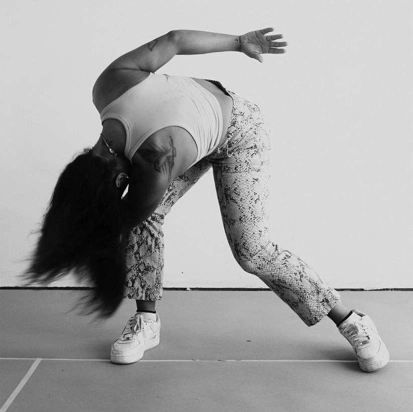 An adult female is bent over, her long dark hair falling towards the ground, wearing a tank top, patterned pants, and sneakers in a monochrome photo.