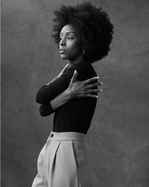 Black and white portrait of a Black woman with an afro, wearing a long-sleeved shirt and pleated trousers, standing in front of a grey backdrop.