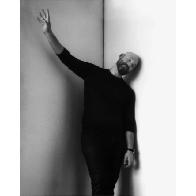 Monochrome studio portrait of a bald adult man with a beard posing with an outstretched arm and hand against a corner wall.