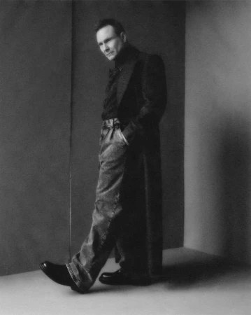 Monochrome full-length portrait of a man in a long dark coat and trousers leaning against a corner.