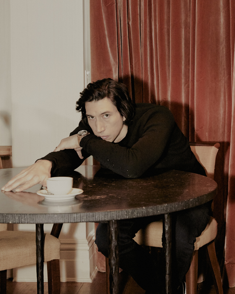 Adam Driver at round table with coffee cup in an indoor scene, wearing a black top with tan and red drapes in the background.