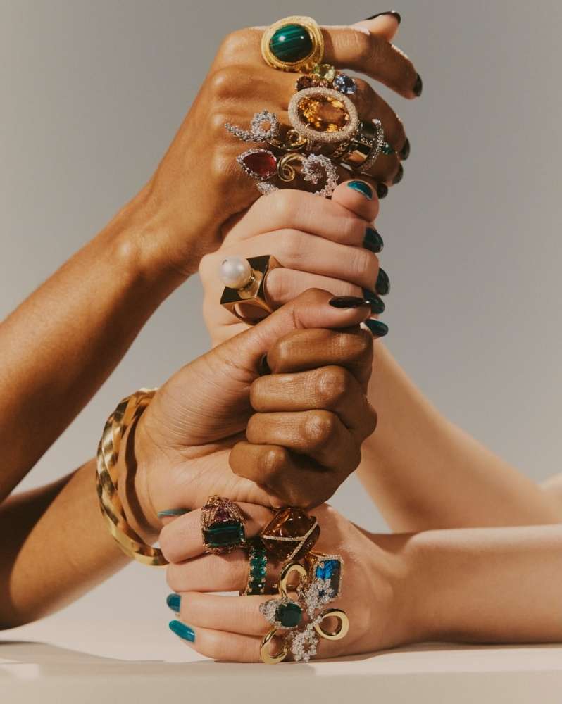 Close-up of three women's hands stacked on top of each other, each wearing multiple ornate rings and a gold bracelet, against a neutral backdrop.