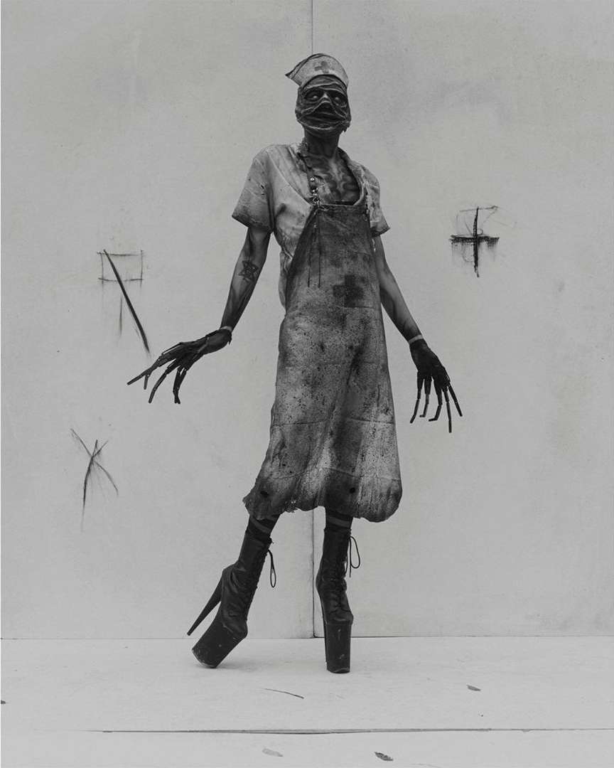 Monochrome image of a nurse-like figure with long fingers and high heels, wearing an apron in front of a white wall.