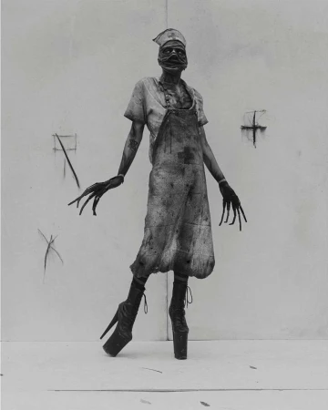 Monochrome image of a nurse-like figure with long fingers and high heels, wearing an apron in front of a white wall.