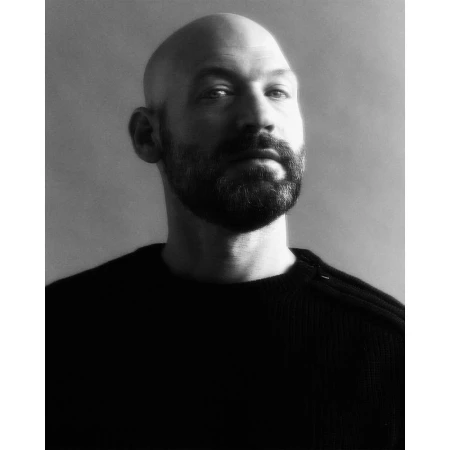 Black and white portrait of a bald adult male with a beard wearing a dark sweater, shown from the chest up against a grey backdrop.