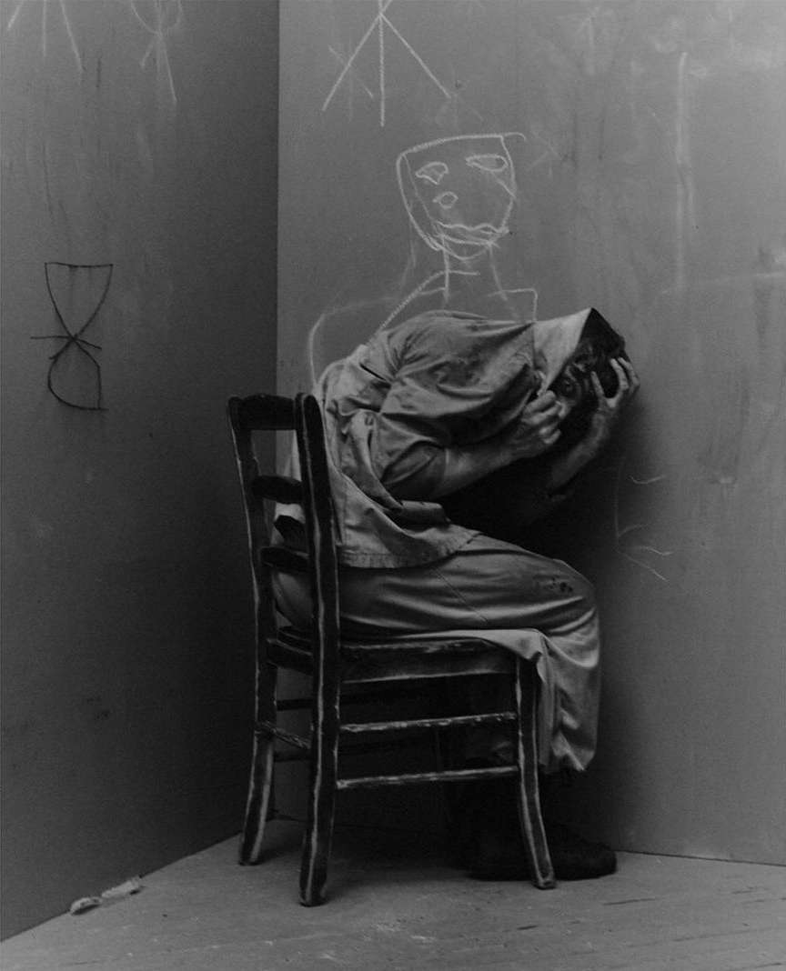 Monochrome image of a person hunched in a chair with chalk drawings on the wall, conveying a mood of distress and introspection.