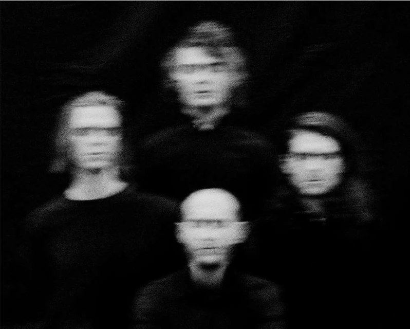 Black and white portrait of a group of four people in darkness, with a shallow depth of field, creating a blurred, vintage aesthetic.