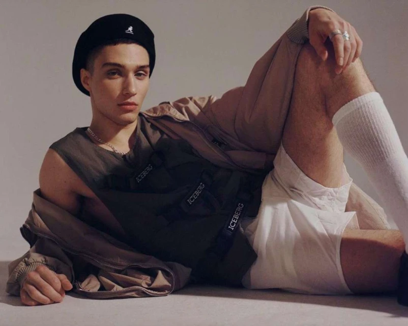 Young adult male model poses while reclining indoors wearing a black beret, olive green vest, white shorts, and white socks.