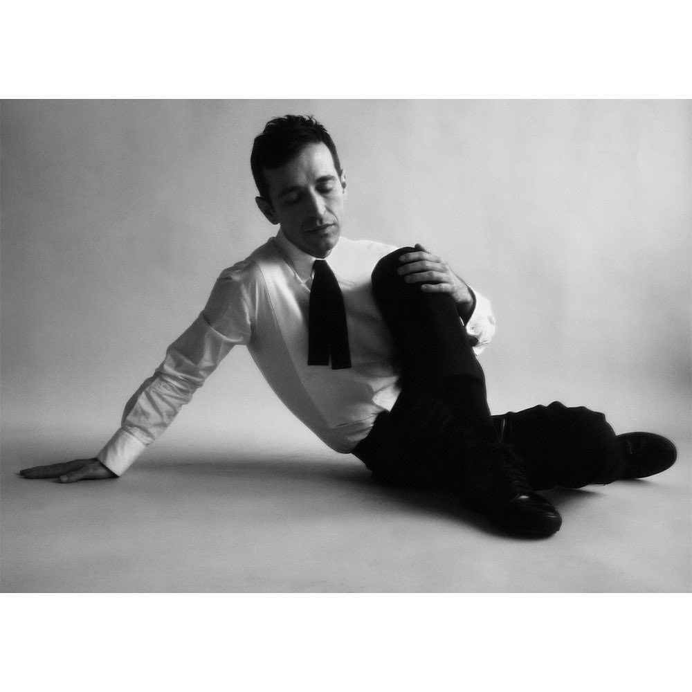 Monochrome portrait of a man in a white shirt and black tie sitting on the floor with one leg bent, striking a contemplative pose.