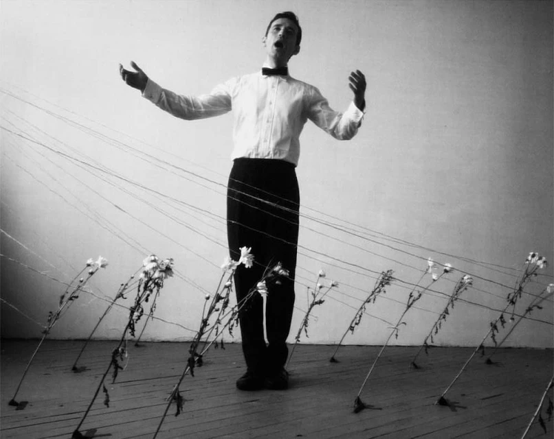 Monochrome portrait of a man singing, wearing a bow tie, surrounded by flowers connected with strings on a wooden floor.