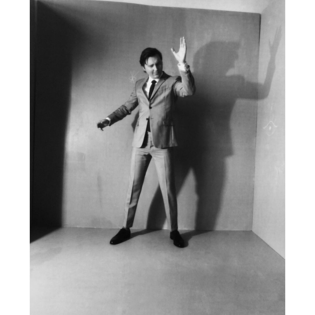 Monochrome studio shot of a man in a suit with raised hand, casting shadow. Gray walls and floor provide a simple, neutral backdrop.