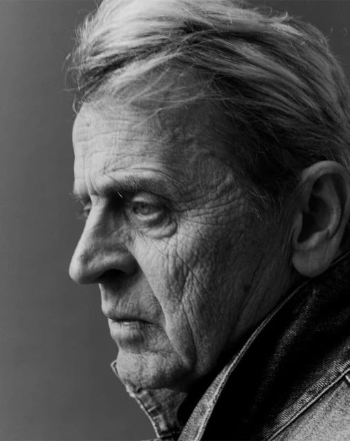 Monochrome portrait of an older adult man in profile, showcasing his textured skin and denim jacket against a solid background.