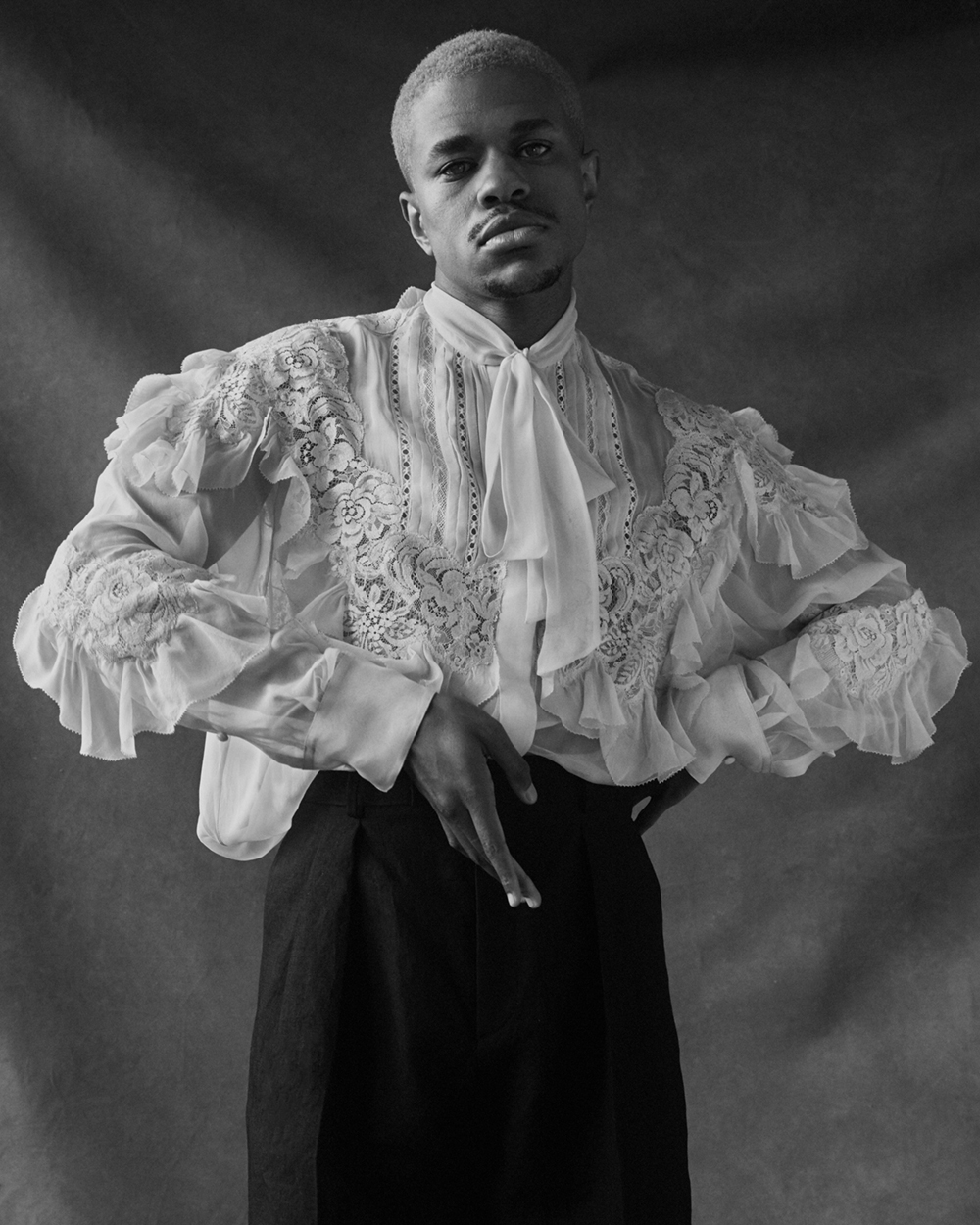 Black and white studio portrait of a fair-skinned Black male model with short bleached hair, wearing a lacy ruffled blouse and dark trousers.