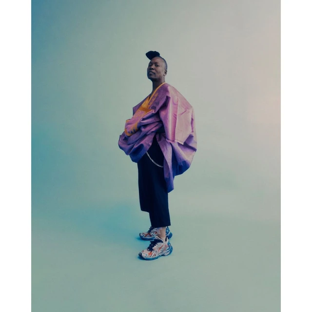 Full-length shot of an adult with a high hairstyle wearing a purple jacket, black pants, and athletic shoes against a light blue background.