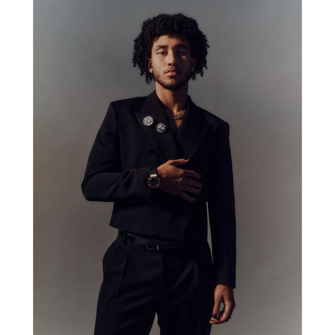 Portrait of a young adult male in a black suit with curly hair posing indoors against a muted backdrop, featuring fashion jewelry details.