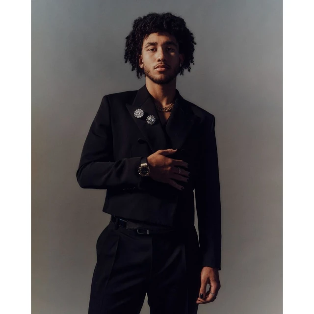 Portrait of a young adult male in a black suit with curly hair posing indoors against a muted backdrop, featuring fashion jewelry details.
