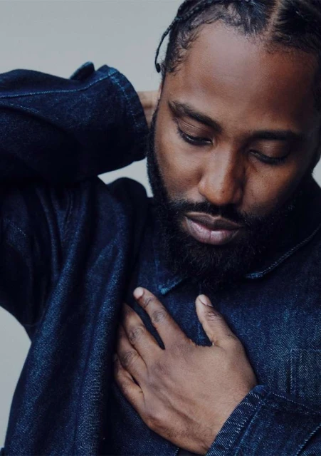 Close-up portrait of a Black man with a beard, wearing a blue denim jacket, posed in an indoor setting with a neutral background.