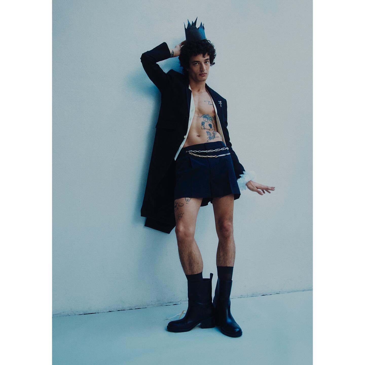 Stylish young man in dark skirt, coat, boots, and crown posing against a wall. Fashion editorial photography, studio lighting, full body shot.