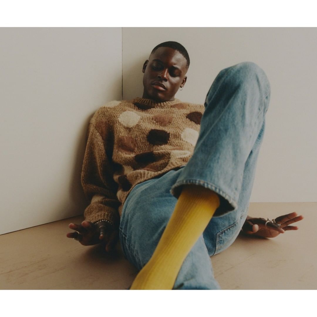 An African-descent adult male poses indoors wearing a brown polka dot sweater, blue jeans, and yellow socks, sitting against a neutral wall.