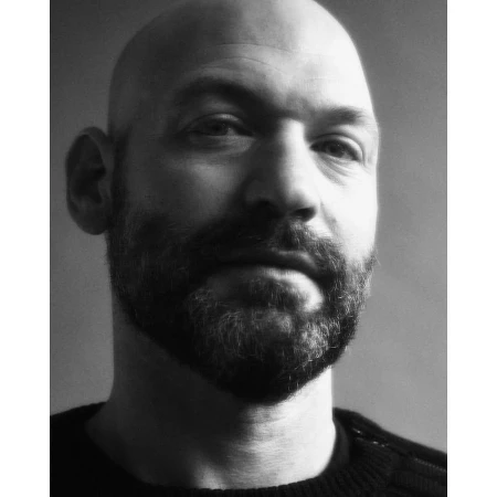 Close-up monochrome portrait of a bald man with a beard wearing a dark sweater against a plain background.