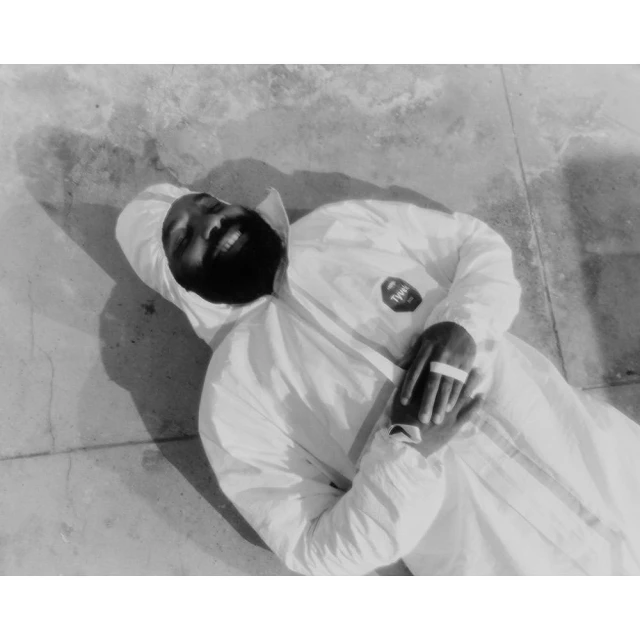 Monochrome portrait of a smiling Black adult male in a white hooded protective suit, lying on a textured gray concrete surface.