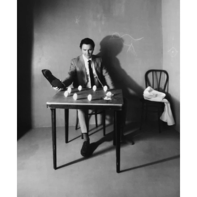 Monochrome studio shot of a man in a suit juggling eggs on a table, holding a shoe, with artistic wall drawings in the background.