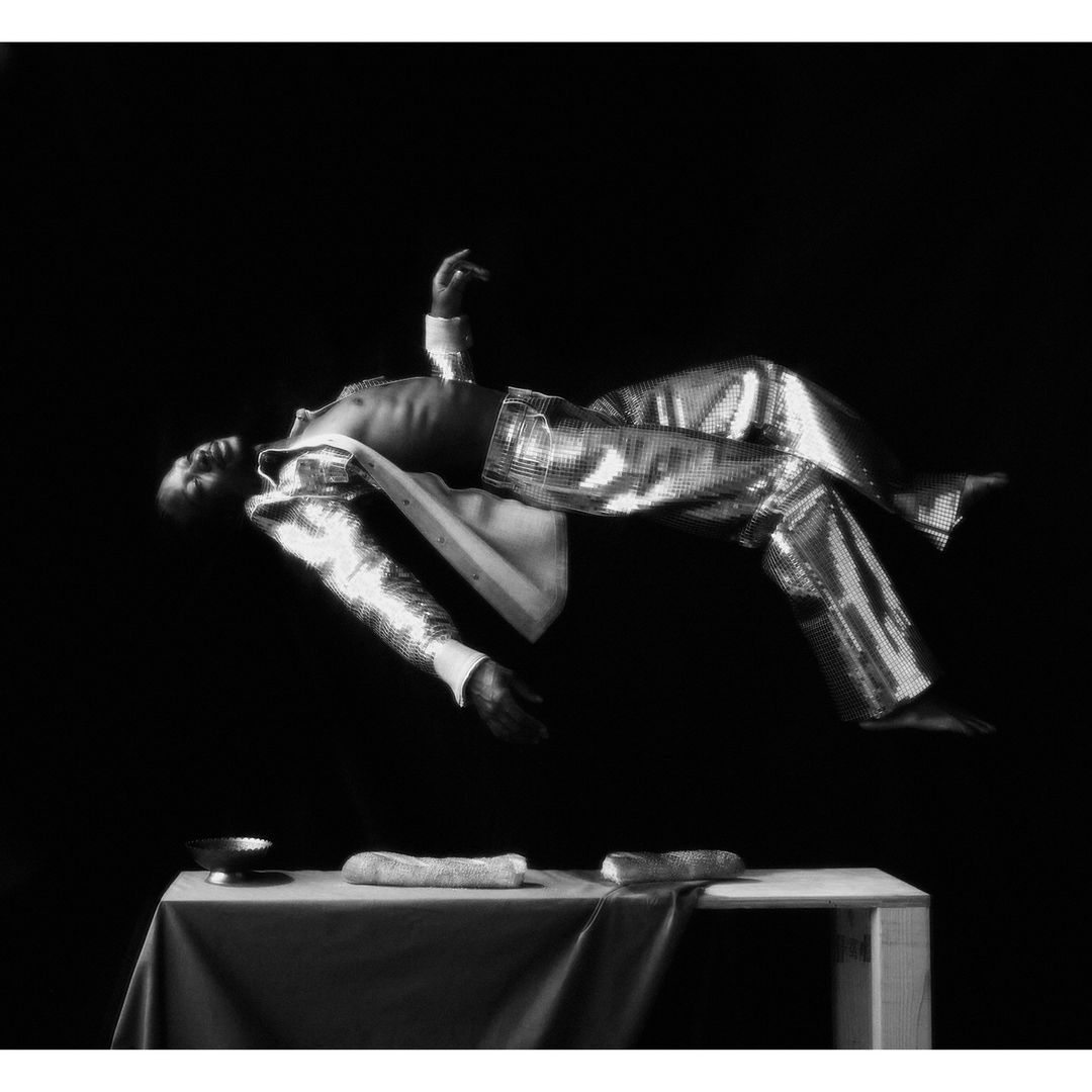 Monochrome artistic studio portrait of a Black man levitating above a table in shimmery pants and jacket against a dark background.