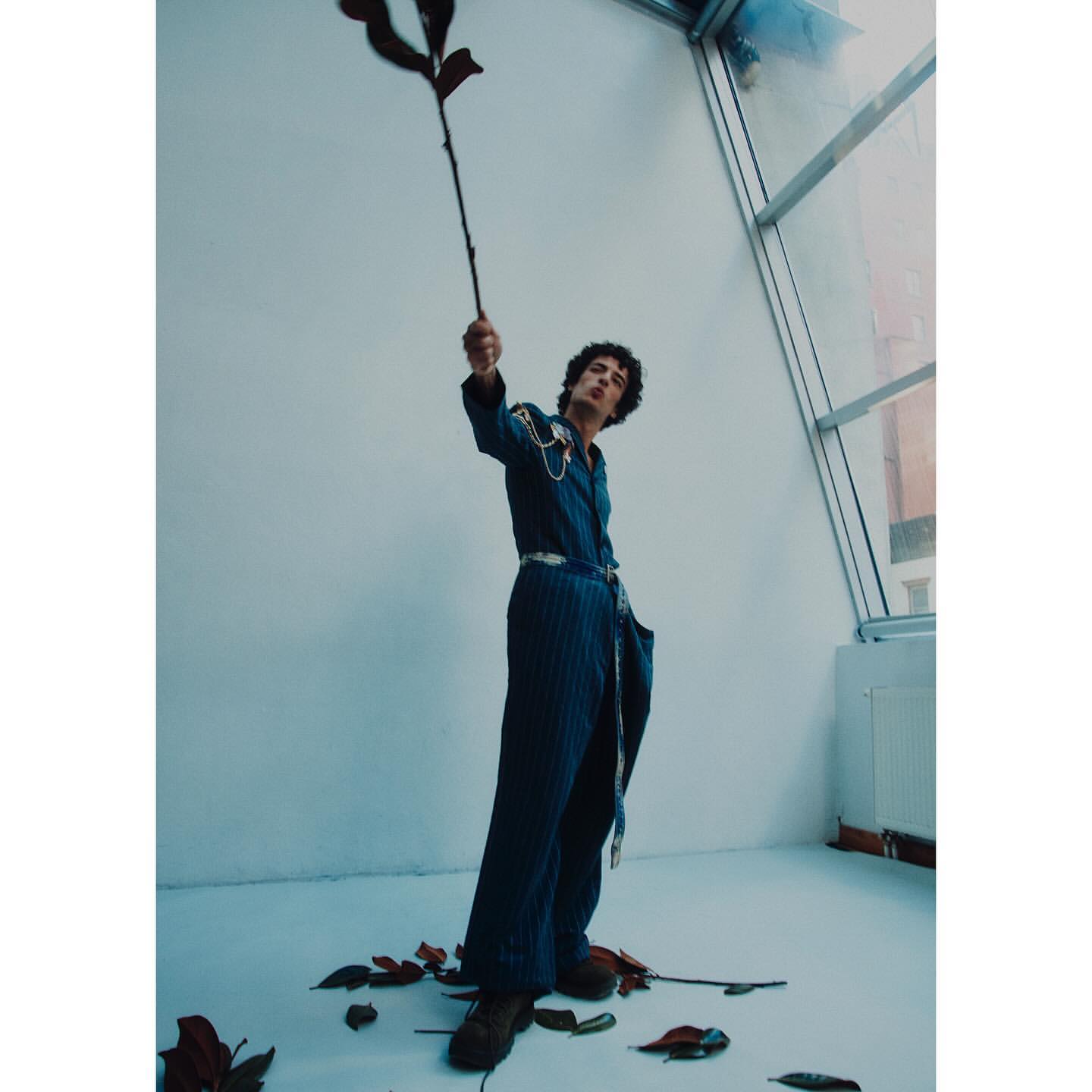 Man in a blue jumpsuit posing in a photo studio, holding a branch with dried leaves, standing near a large window with city view.